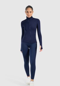 Chaqueta deportiva azul marino con cuello alto, cremallera completa y diseño entallado. Combinada con leggings a juego, que presentan una textura suave y sin patrones.