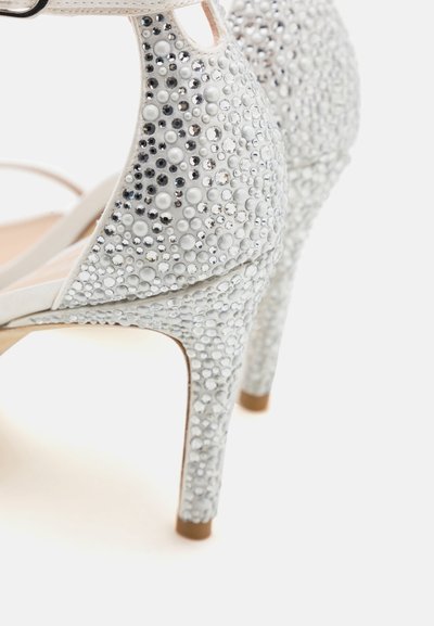 Sandale à talons blanche avec une texture lisse, ornée de gemmes circulaires et de strass, dotée d'une bride de cheville et d'un design à bout pointu.