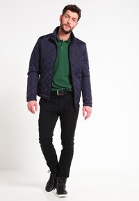 Chaqueta de náutica acolchada con cierre, llevada sobre una camisa polo verde. Combinada con pantalones negros y zapatillas de caña alta negras.