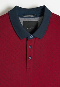 Koszulka polo o dopasowanym kroju w kolorze bordowym z granatowym kołnierzem, ozdobiona małymi ciemnymi wzorami, z trzema guzikami i metką wewnątrz z napisem „diverse”.