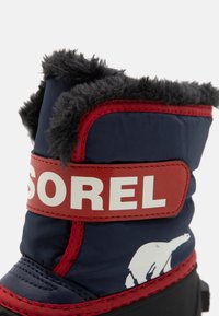 Marineblaue und rote Winterstiefel mit Kunstfellbesatz, Sorel-Logo-Riemen und einem Eisbären-Motiv an der Seite; wasserdichtes Material und warme Fütterung.