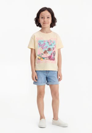 Barn med skulderlangt hår iført en gul Puerto Rico grafisk t-shirt, denimshorts og hvide slip-on sko, står på hvid baggrund.