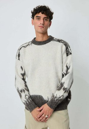 Jeune homme aux cheveux bouclés portant un pull en tricot blanc et gris foncé à motifs et un pantalon beige, se tenant devant un fond uni.