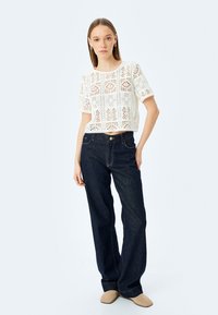 Top corto de encaje blanco con mangas cortas, combinado con jeans de mezclilla azul oscuro de tiro alto. La modelo lleva zapatos de tela color beige.