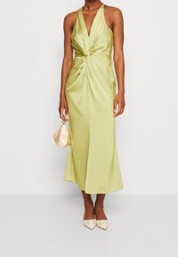 Žena v bezrukávých světle zelených saténových midi šatech se zkrouceným předním dílem, drží malou béžovou saténovou kabelku a béžové špičaté boty.