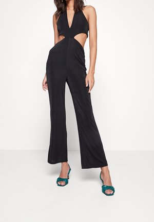 Czarny kombinezon z głębokim dekoltem w kształcie litery V, wycięciami po bokach i rozszerzanymi nogawkami. Wykonany z gładkiej tkaniny. Zestawiony z tealowymi sandałami na obcasie z odkrytymi palcami.