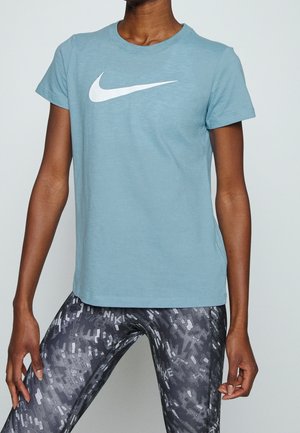 Personne portant un t-shirt Nike bleu clair avec un logo blanc et des leggings Nike noirs à motifs sur un fond uni.