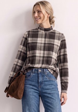 Kvinde smiler, mens hun har en brun og beige ternet langærmet top, blå jeans og holder en brun ruskindstaske.