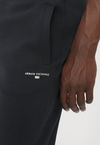Black jogger pants made of cotton blend, featuring a side pocket and a white embroidered logo "ARMANI EXCHANGE 1991" on the thigh.