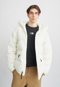 Doudoune blanche avec capuche, dotée d'une fermeture éclair à l'avant, de deux poches et d'accents noirs. Portée sur un t-shirt noir et un pantalon beige.
