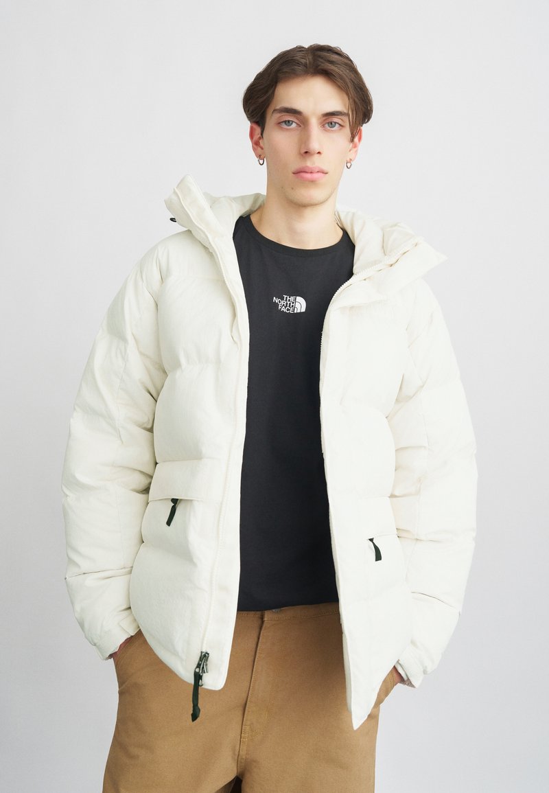 Doudoune blanche avec capuche, dotée d'une fermeture éclair à l'avant, de deux poches et d'accents noirs. Portée sur un t-shirt noir et un pantalon beige.