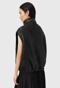Giacca senza maniche in pelle nera con colletto alto. Texture liscia, vita elastica e design aderente. Dettagli con zip sul collo.