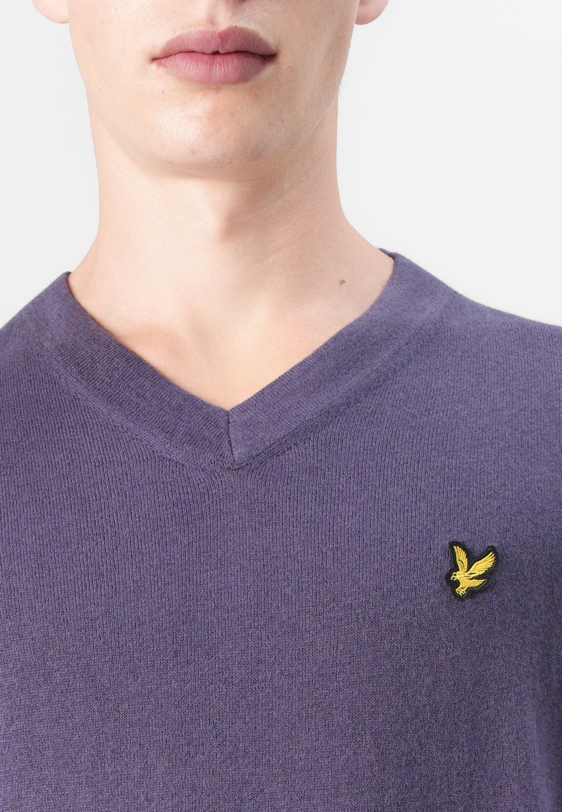 Sweter robiony na drutach w kolorze fioletowym z dekoltem w kształcie litery V, wyróżniający się teksturowanym materiałem. Mała żółta haftowana logo ptaka znajduje się po lewej stronie klatki piersiowej.