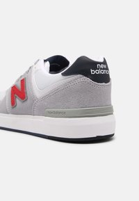 Šedé, bílé a námořnické tenisky New Balance s červeným logem "N", vyrobené z semiše a síťoviny, viditelné značení na patě, šikmý pohled na bílém pozadí.