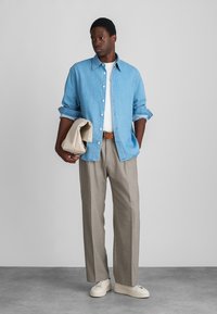 Lichtblauwe overhemdd, wit t-shirt, beige broek, houdt een lichtgekleurde tas vast en draagt witte sneakers. Eenvoudige, relaxte silhouet.