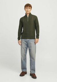 Dunkelgrüner Strickpullover mit Reißverschlusskragen, der strukturierte Zopfmuster aufweist. Kombiniert mit hellblauen Jeans und braunen Schuhen.