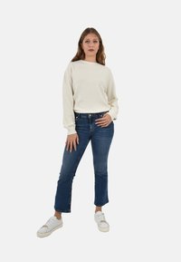 Maglione bianco a trama abbinato a jeans svasati blu e sneakers bianche, caratterizzato da una vestibilità casual e un design minimale, in piedi davanti a uno sfondo bianco.