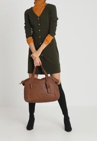 Bolso de piel marrón con acentos de borlas, de forma rectangular y con asas dobles. La modelo lleva un vestido verde y calcetines negros.