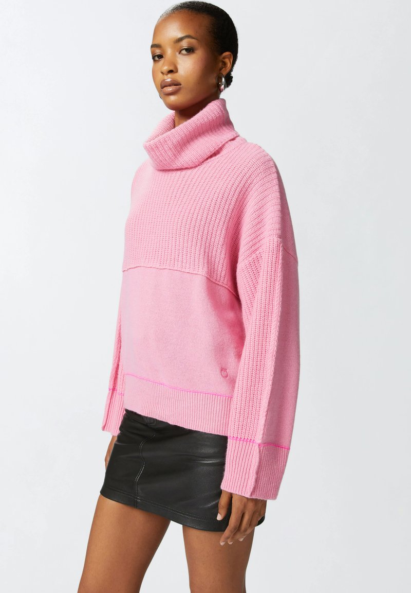 Pull en tricot rose surdimensionné avec un col roulé haut, une texture côtelée et une partie inférieure contrastante. Associé à une mini-jupe en cuir noir.