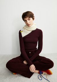 Rippenshirt in Bordeauxrot mit langen Ärmeln, getragen zu dunkelroten Cordhosen. Weicher cremefarbener Schal, der um den Hals drapiert ist, mit einem Etikett und einem kontrastierenden gelben Akzent.