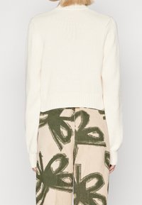 Person med kort blont hår som står vänd bort, iförd en krämfärgad stickad tröja och beige byxor med stora mörkgröna abstrakta blommönster.