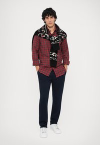 Czerwona i czarna koszula w kratę noszona pod czarnym, wzorzystym, dzianinowym szalem, granatowe spodnie i białe trampki. Prosty i casualowy design.