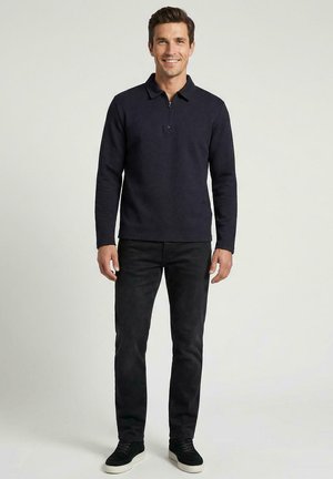 Man staat en lacht, draagt een donkerblauwe trui met lange mouwen en rits, zwarte broek en zwarte sneakers met witte zolen tegen een effen achtergrond.