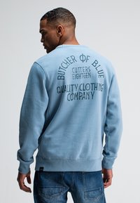 Lichtblauwe sweatshirt met gedurfde tekst in de rug. Gemaakt van zachte stof, heeft het ribgebreide manchetten en een ronde hals. Gepaard met denim jeans.