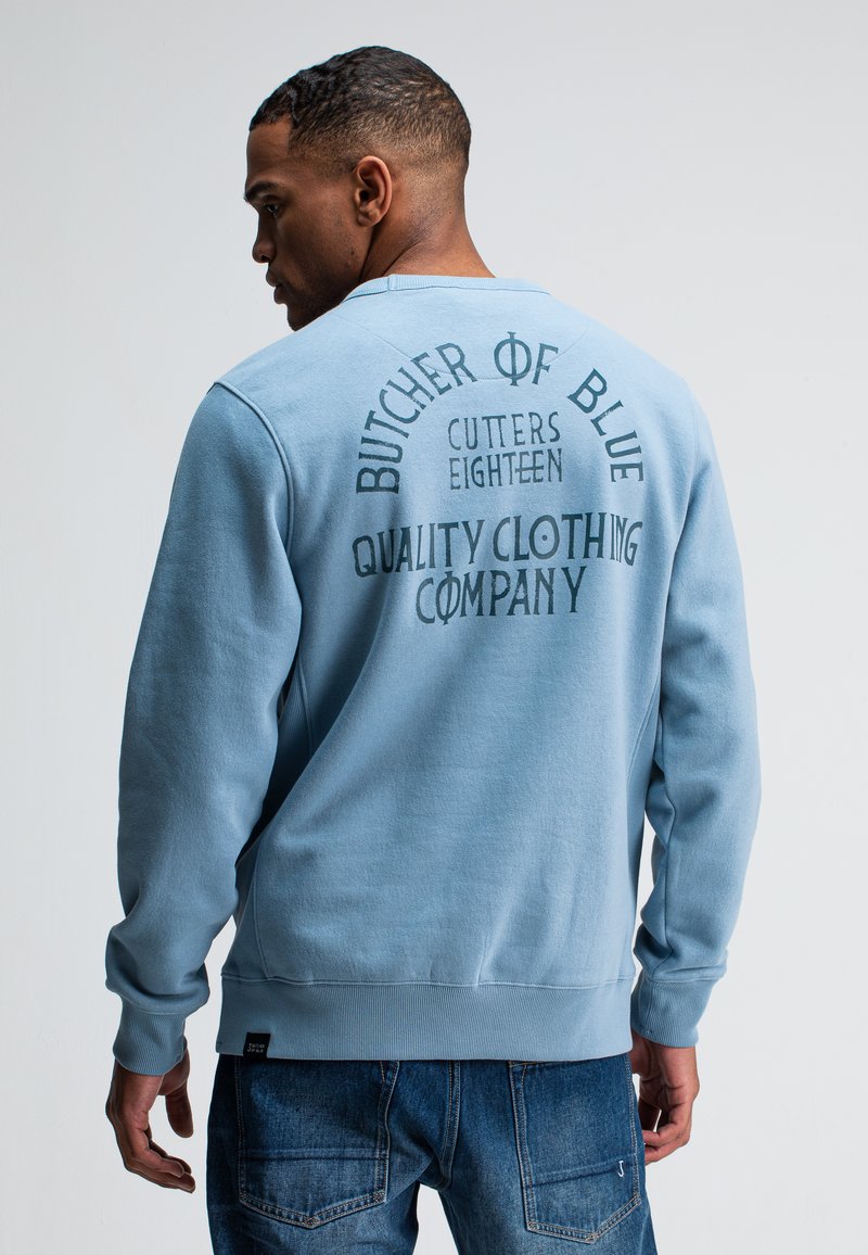 Lichtblauwe sweatshirt met gedurfde tekst in de rug. Gemaakt van zachte stof, heeft het ribgebreide manchetten en een ronde hals. Gepaard met denim jeans.