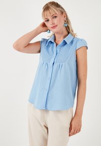 Blusa azzurra chiara con maniche corte, chiusura a bottoni e dettagli in pizzo sul davanti. Tessuto morbido con vestibilità rilassata.