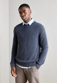 Strikket marineblå genser med ribbet tekstur, rund hals, brukt over en lyseblå krageskjorte, kombinert med beige bukser.