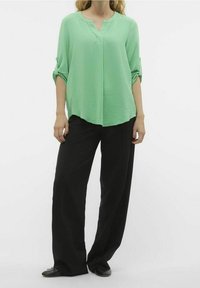 Mátově zelená blouse s výstřihem do V a ohrnutými rukávy, spárovaná s volnými černými kalhotami a černými botami. Hladká látka se slabou texturou.