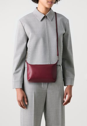 Sac en bandoulière en cuir bordeaux avec une texture lisse, de forme rectangulaire et une sangle ajustable, porté avec une tenue à carreaux noir et blanc.