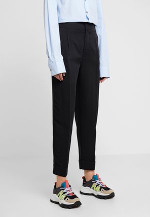 Personne portant un pantalon noir à revers, une chemise bleu clair à manches longues et des baskets épaisses multicolores, debout sur un fond uni.