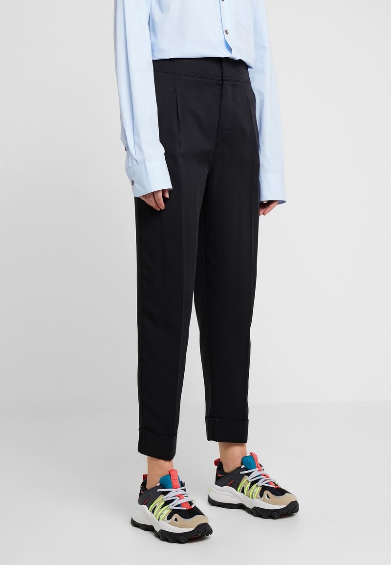 Personne portant un pantalon noir à revers, une chemise bleu clair à manches longues et des baskets épaisses multicolores, debout sur un fond uni.