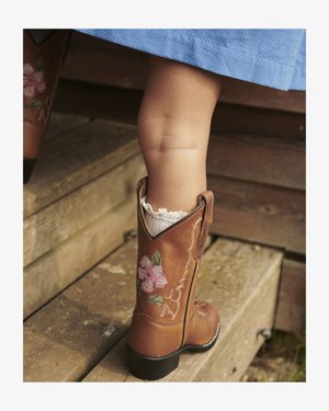 Beenen van een kind met een bruine leren cowboylaars met roze bloemrijk borduurwerk en een witte sok met kanten rand, die op houten trappen stapt.