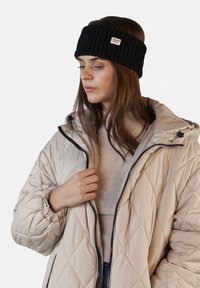 Fascia nera in maglia con texture rigata e una piccola etichetta con logo, abbinata a una giacca trapuntata beige e a un maglione chiaro sottostante.