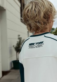 Biely mikina s zelenými akcentmi, na prednej strane s čiernym potlačeným textom "NEW YORK" a na zadnej strane s názvami piatich štvrtí. Mäkká tkanina, uvoľnený strih.