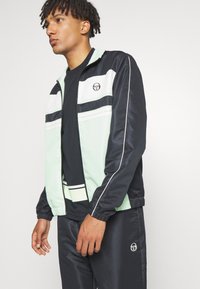 Juoda ir mėtų žalia striukė, su elastiniais rankogaliais, logotipu ant krūtinės ir dryžuotomis detalėmis. Suderinta su atitinkančiomis juodomis kelnėmis.