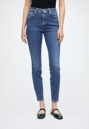 Femme portant un jean skinny bleu taille moyenne et des chaussures Mary Jane noires, debout sur un fond blanc uni.