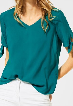 Frau trägt eine türkisfarbene V-Ausschnitt-Bluse mit kurzen Ärmeln, die zu Schleifen gebunden sind, kombiniert mit weißen Hosen, gezeigt vom Hals bis zur Hüfte.