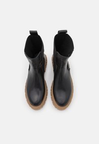 Paire de bottines en cuir noir avec semelles épaisses en caoutchouc marron clair, languettes à l'arrière, vues de dessus sur fond blanc.