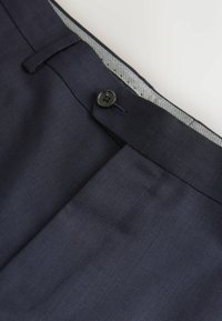 Navy trousers featuring a flat front design, front pocket, and a black button closure. Interior has a striped fabric accent on the waistband.