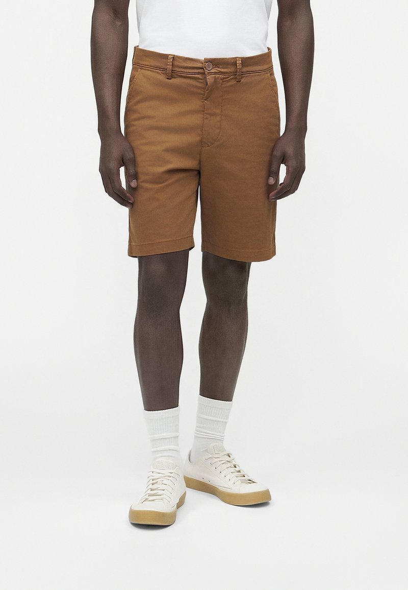 Άνδρας που φοράει καφέ βερμούδα μέχρι το γόνατο, λευκές κάλτσες και λευκά casual αθλητικά παπούτσια, στέκεται μπροστά σε απλό φόντο.