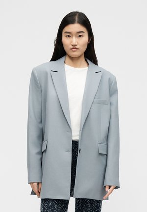 Femeie cu păr lung și închis la culoare, purtând un sacou supradimensionat gri deschis peste un top alb și pantaloni în pattern închis, pe un fundal simplu.