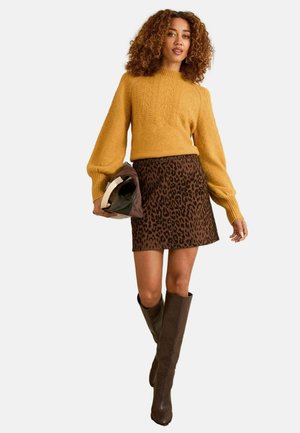 Senf gestrickter Pullover mit komplexem Muster, brauter Leopardenmusterrock und knielangen braunen Stiefeln, hält eine braune Clutch-Tasche.