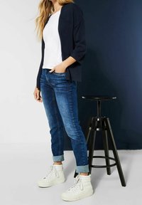 Laivastonsininen neulottu neulejakku, valkoinen pyöreäkauluksinen t-paita, siniset slim fit -farkut ja valkoiset korkeavartiset tennarit. Taustalla musta puinen jakkara.