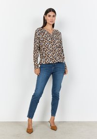 Langärmlige gemusterte Bluse mit braunen, weißen und blauen abstrakten Designs. Kombiniert mit eng anliegenden blauen Jeans und hellbraunen spitzen High Heels.