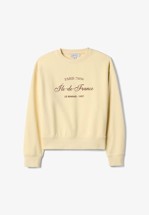 Sudadera amarilla con cuello redondo, texto impreso en marrón: "PARÍS 75056 Île-de-France LE MARAIS - 1997." Puños y dobladillo acanalados.