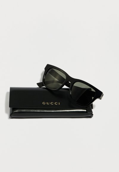 Γυαλιά ηλίου με μαύρο τετράγωνο σκελετό, τοποθετημένα πάνω σε μαύρη θήκη Gucci με χρυσό λογότυπο, σε λευκό φόντο.